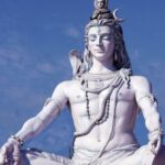 A tall statue of Lord Shiva, who sits with his legs crossed and eyes closed. The status is white and Shiva wears pants, with a snake draped around his neck along with other symbolic adornments.