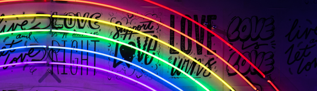 Neon lights in a rainbow shape and color against a wall with affirmations of love in various text shapes: Live and Let Love, Love is a Right, Support Love, Love Wins, Love is Love. By photographer Jason Leung, free license from Unsplash.com. 