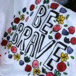 White cloth with with colorful painted flowers surrounding text "be brave."