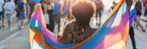 Two people with brown skin holding a rainbow colored scarf around their shoulders walking with a crowd waving rainbow flags.