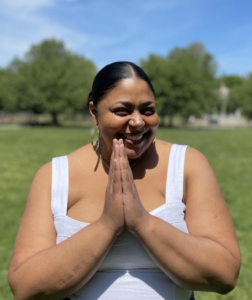 Elisanett Martinez smiles, palms together, with a field of grass and trees in the background.