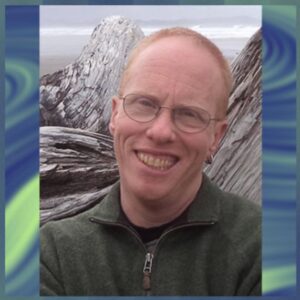 Eli Clare smiles at camera wearing a green zip up top, classes, and hoop earrings in right ear, with gray rocks and cloudy sky in background