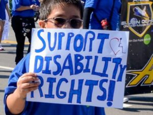 A child takes part in the Los Angeles Disability Pride Parade, holding a sign with blue lettering "Support Disability Rights."