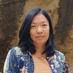 Susie Ibarra wearing a tan shirt and floral overshirt, standing in front a rocky backdrop.