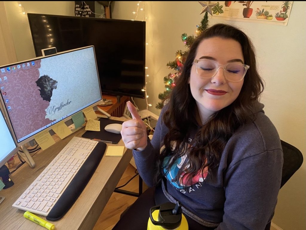 Hanna smiles and gives a thumbs-up to the camera while working on class projects in front of her computer. 