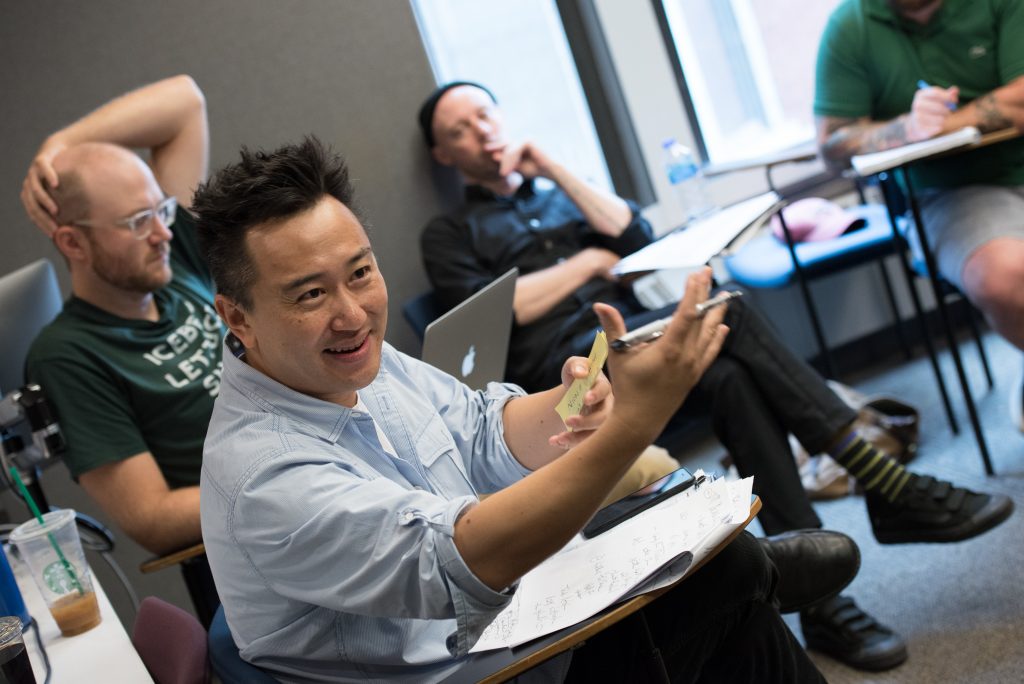 Emerson professor sitting at a classroom desk, gesturing animatedly with their hands to students in a writing workshop, three students listening and taking notes in the background.