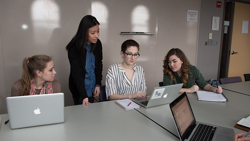 Four graduate students collaborating on a project, all looking at one student's laptop screen on the table in front of them.