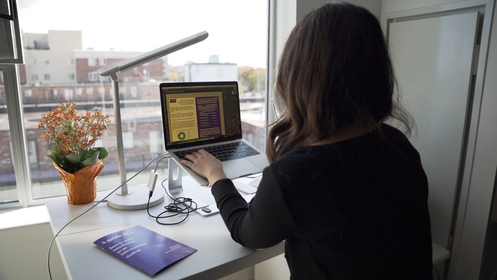 Shot of Media Design student working on the computer at her desk. Taken from behind her to show her view out the window behind her computer. The completed purple zine is next to her on the desk. 