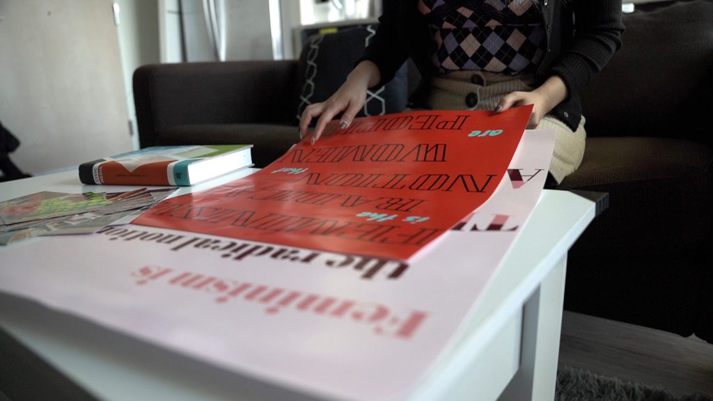 Sharry's Media Design posters on the coffee table: red background with black all-caps text that reads: "Feminism is the radical notion that women are people." 