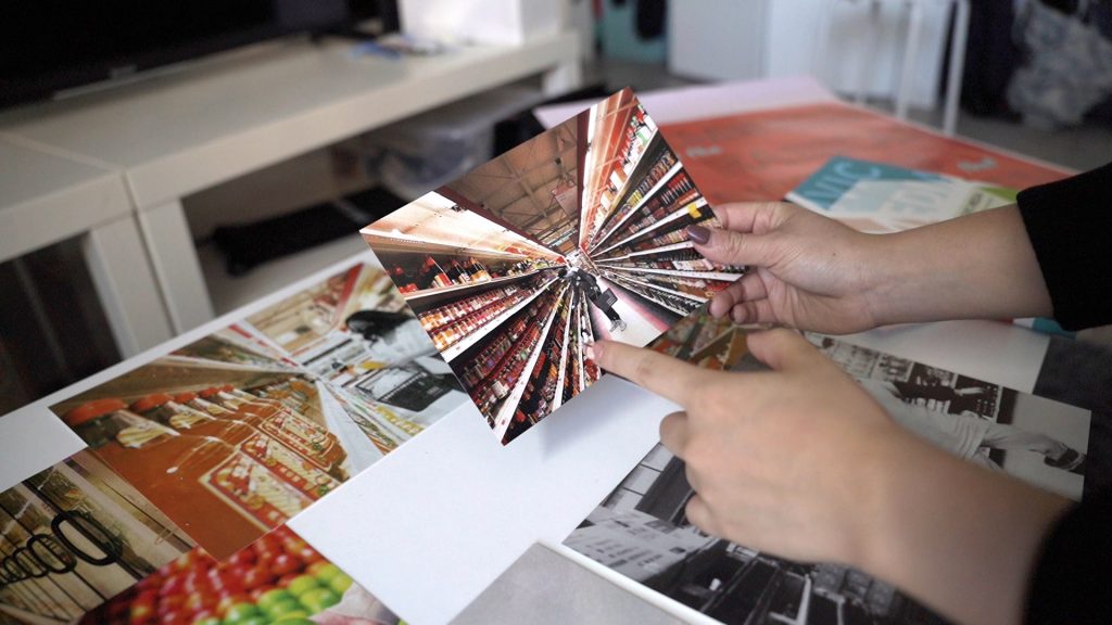 Several printed photos from Sharry's Media Design Hong Kong project, some laying on the table, one of them held in her hands, featuring people shopping in the local Super88 grocery store. The people in the images have been de-colorized in black and white while the background colors of the aisles have been saturated with color to create a contrasting effect. The image Sharry is holding features a person standing in a narrow aisle, considering the products to their left.