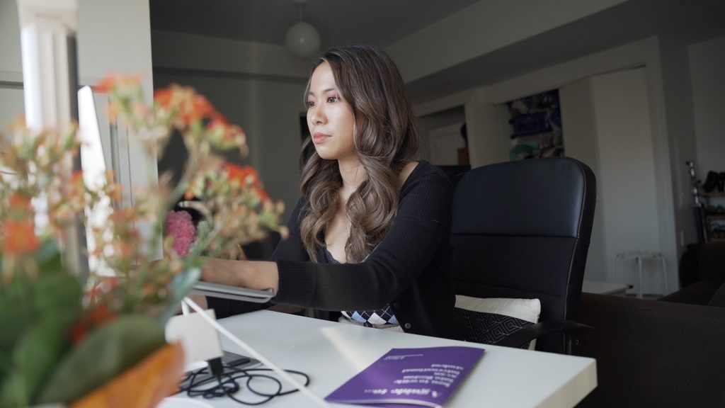 Sharry Li sitting at her desk and working at her desktop computer, her Zine "How to Settle into Boston as an International Student" with a purple cover and white text on the desk next to her.