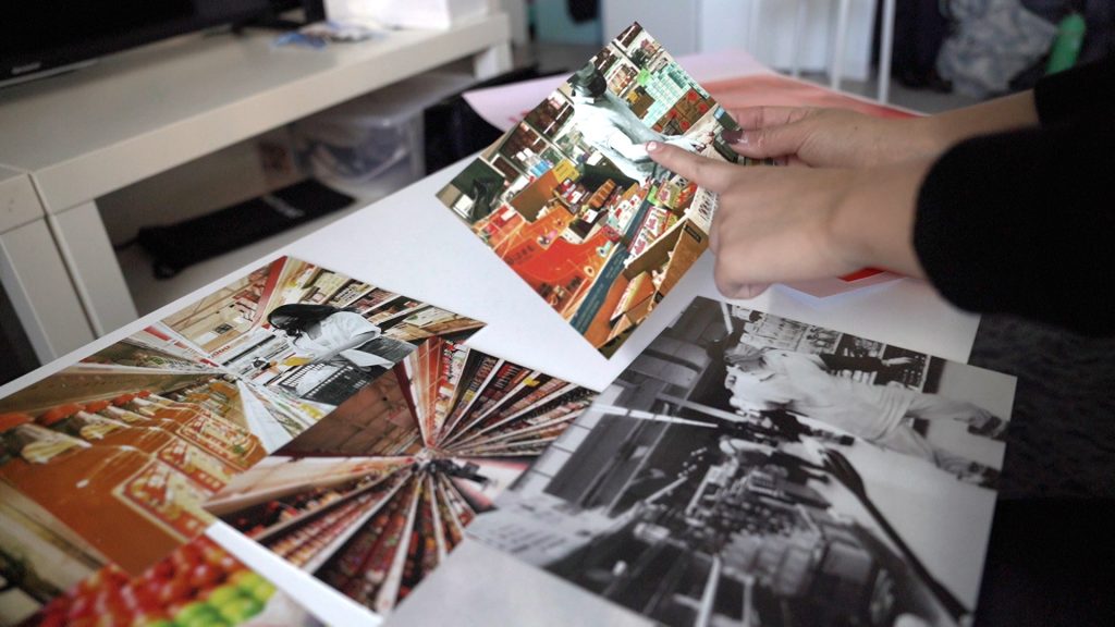 Several printed photos from Sharry's Hong Kong project for Media Design. Photos are laying on the table, one of them held in her hands, featuring people shopping in the local Super88 grocery store. The people in the images have been de-colorized in black and white while the background colors of the aisles have been saturated with color to create a contrasting effect. The image she is holding features a masked man selecting a product from a bin in front of him.