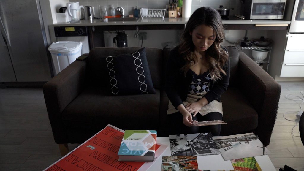 Sharry sitting on a couch in her apartment, looking at her collection of work (posters and photos) and a textbook from the Media Design program on the coffee table in front of her.