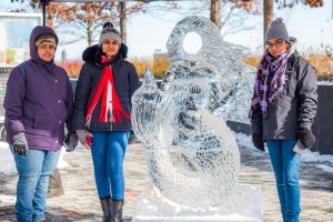 Three women smiling next to a waterfront ice sculpture 