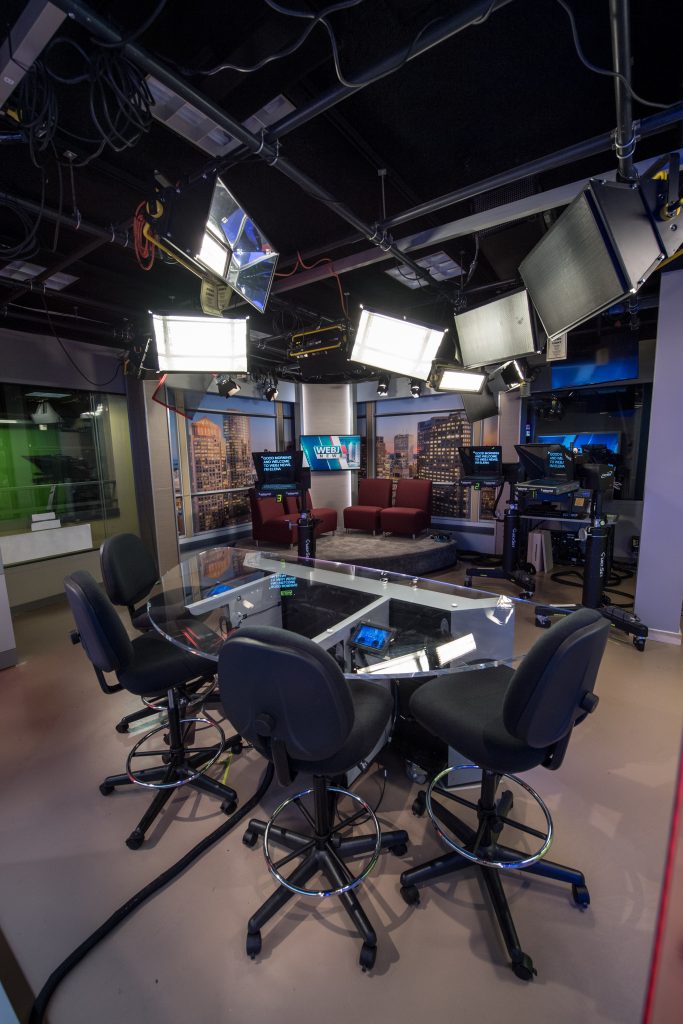 A journalism broadcasting room is set up with lights and a table and chairs for journalism students to practice in.