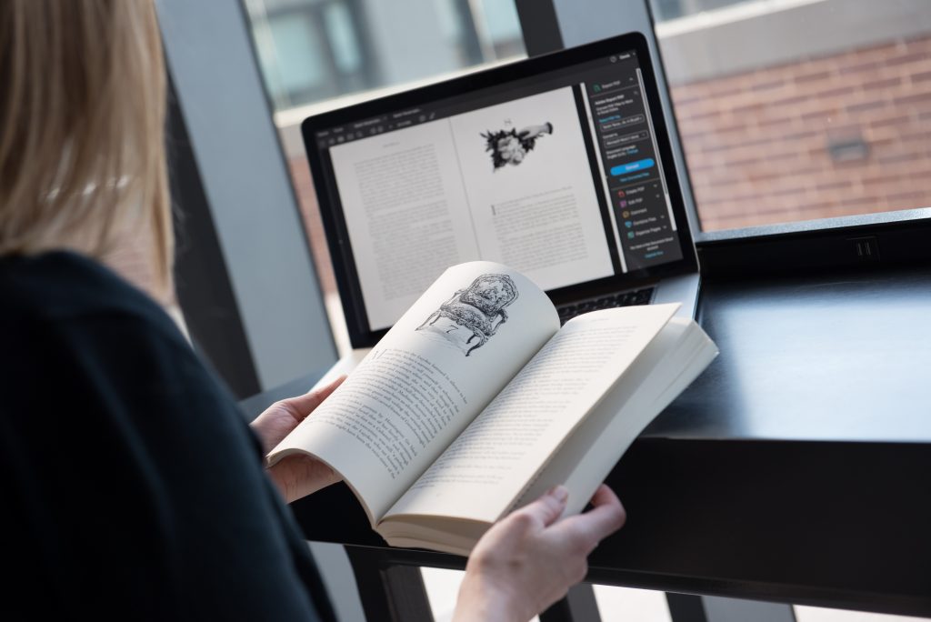 A student works on book design with their laptop open behind the book they are holding. 