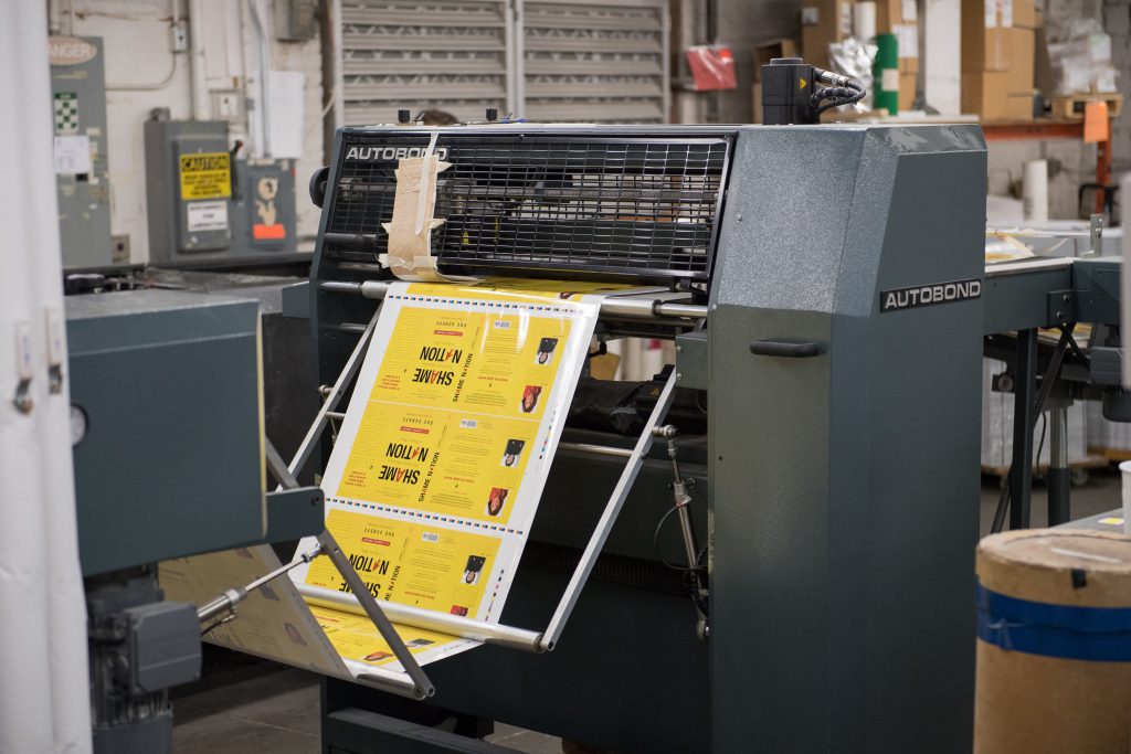 A printing press in action, printing several copies of a book cover on bright yellow paper at a local Boston publishing house as part of a tour for a course in Book Design.