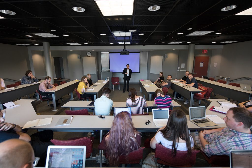 A graduate writing course in action, the professor stands at the front of a classroom where students are seated at long tables with laptops or notebooks in front of them. 
