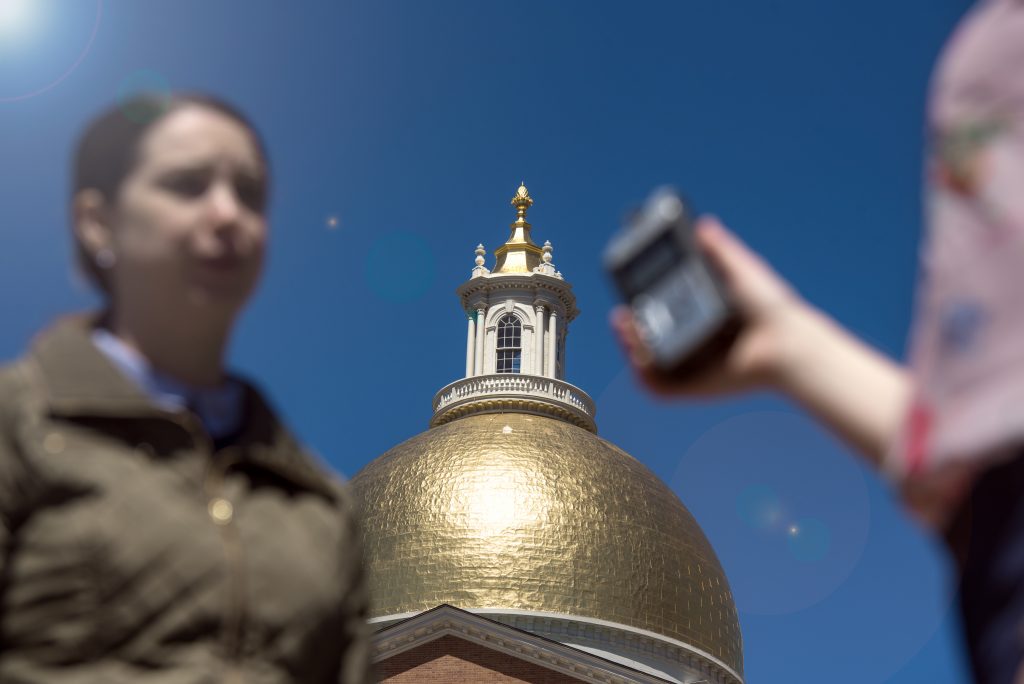 An Emerson graduate student interviews an individual in front of the state house, image shows student holding the recording device while the interviewee speaks. 