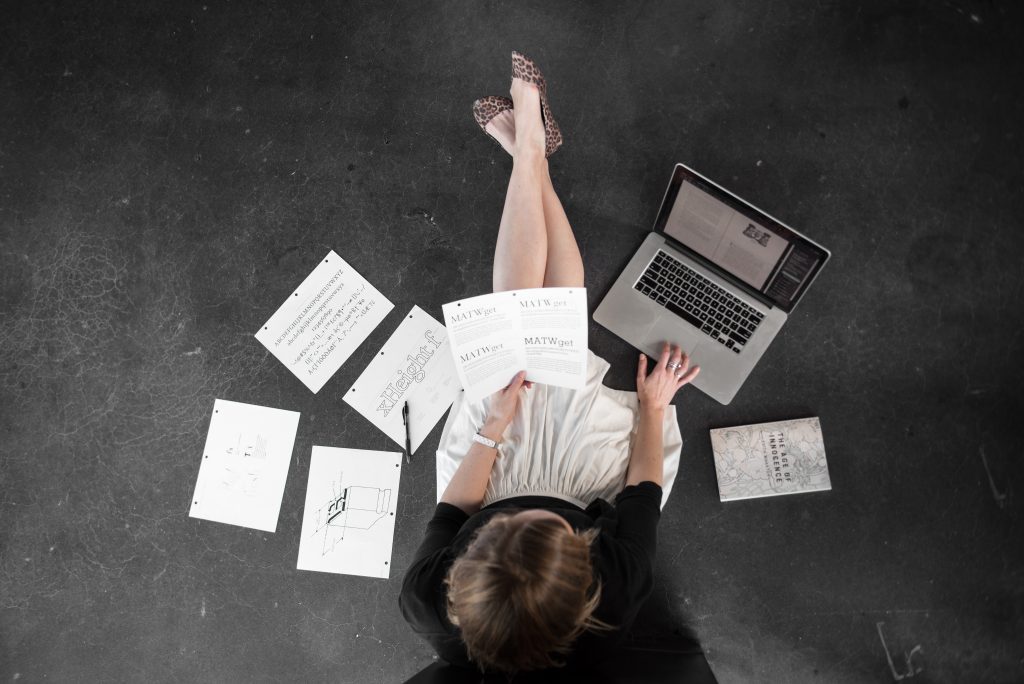 Grad student with book design materials spread around them, sitting on the floor, working on a laptop.