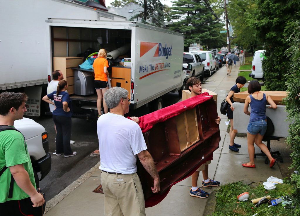 Two groups of people carrying couches on the street, coming in and going out of a driveway, moving trucks in the background. Moving weekend in Allston, Photo from Boston WBUR 