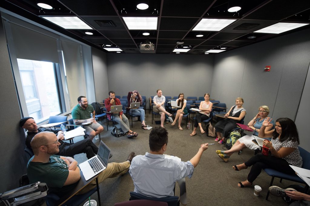 Image of a Creative Writing MFA workshop in action, 11 students and the professor seated at desks facing each other in a circle, the professor in the center of the photo, pointing at a student with a raised hand to their right.