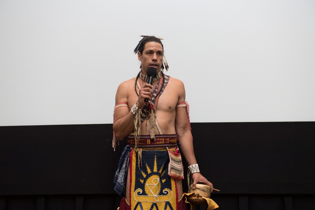 Man dressed in traditional Wampanoag clothing speaks into a microphone
