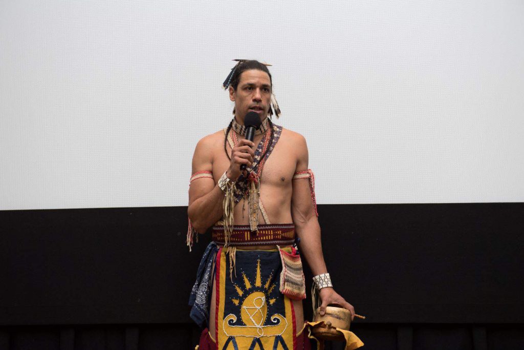 Man dressed in traditional Wampanoag  clothing speaks into a microphone