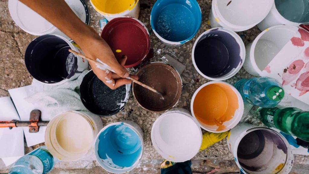 An artist hovers their brush over large pails of colorful paint