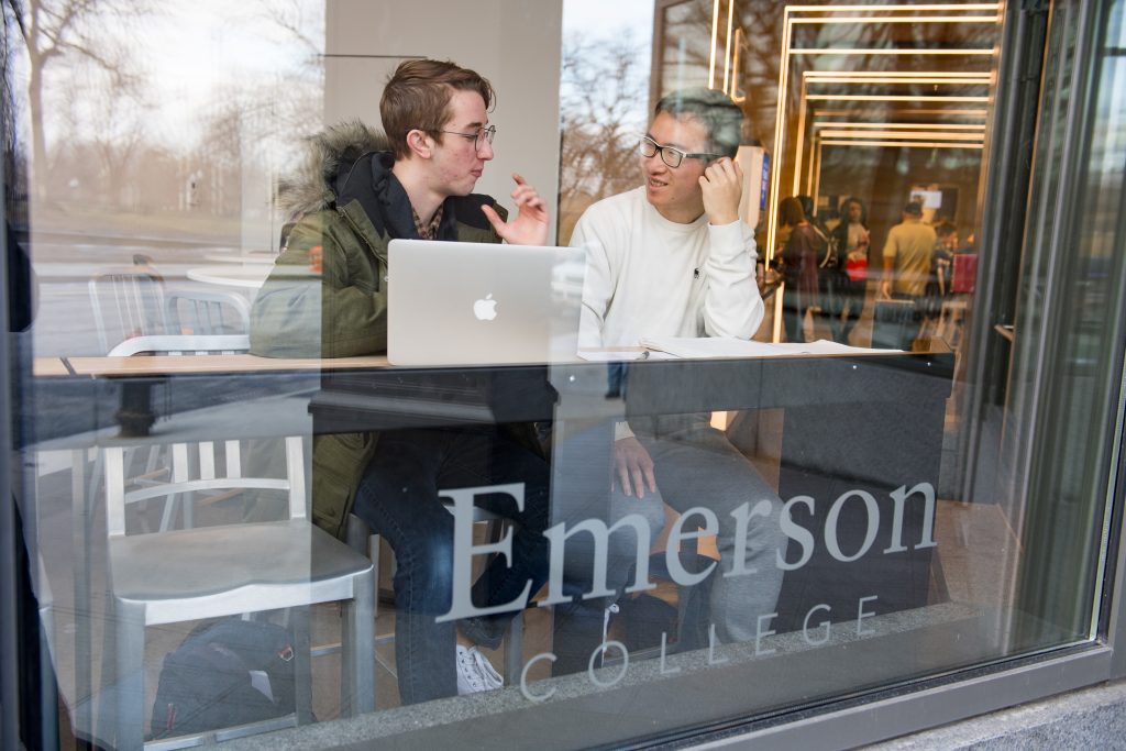 Two Emerson students sit talking behind a window with "Emerson College" etched into the glass.