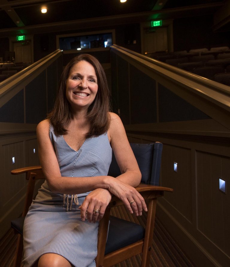 Laura Gallagher Byrne sitting in a chair in a dimly lit theater, smiling at the camera
