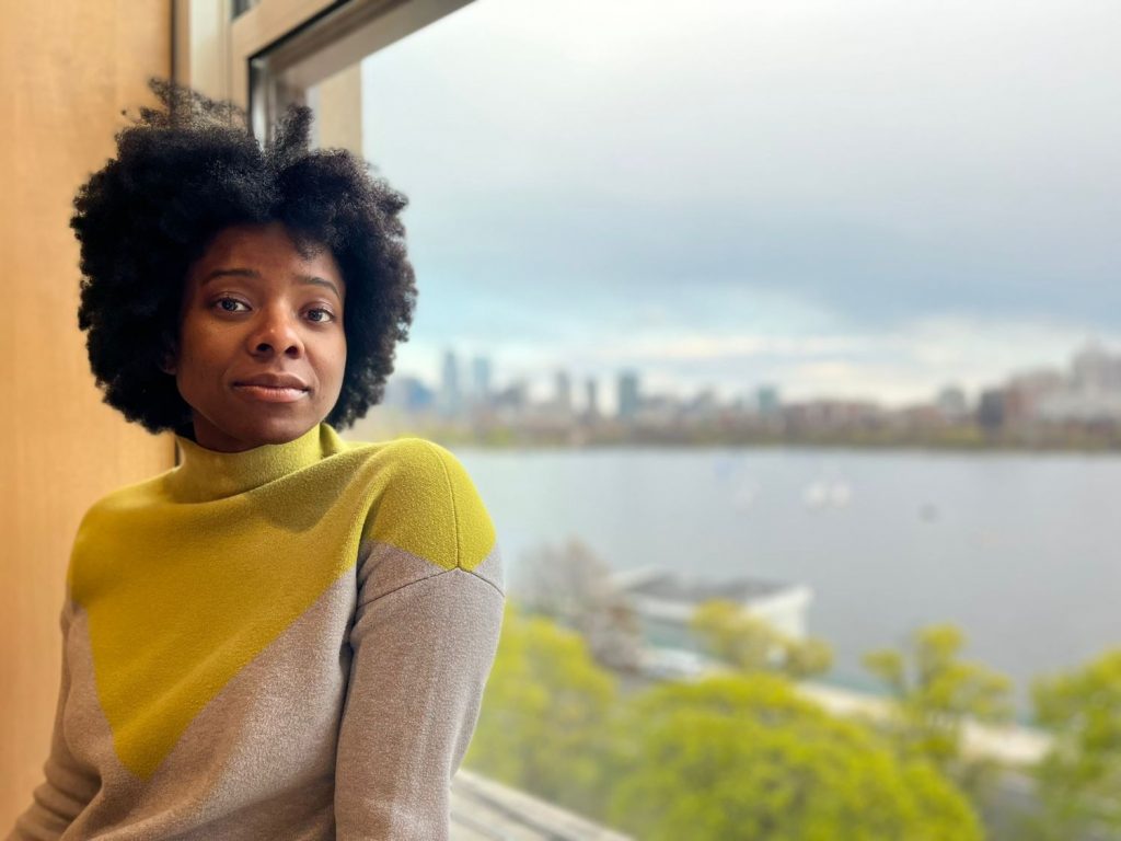 Dom Lounds in a yellow and grey sweater with the Boston skyline behind her