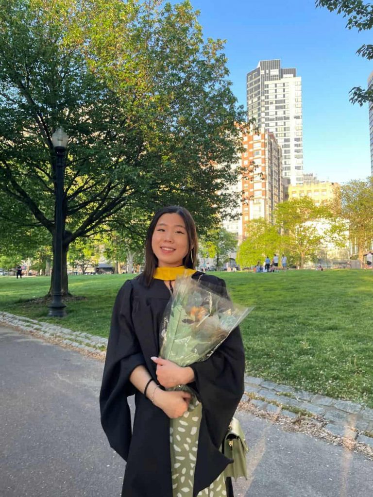Alisha Lai standing in the Boston Common in a graduation robe and holding flowers