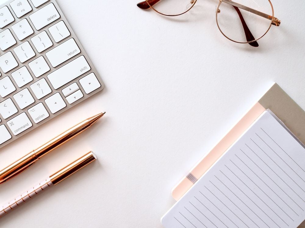 Glasses, a computer keyboard, pens, and notepads laying on a desk