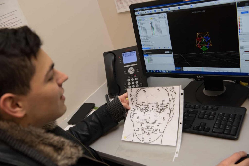 A Commuication Disorders student completing research work in one of the faculty's research labs. The student sits at a desktop computer and holds a drawing of a face with different points of the face highlighted