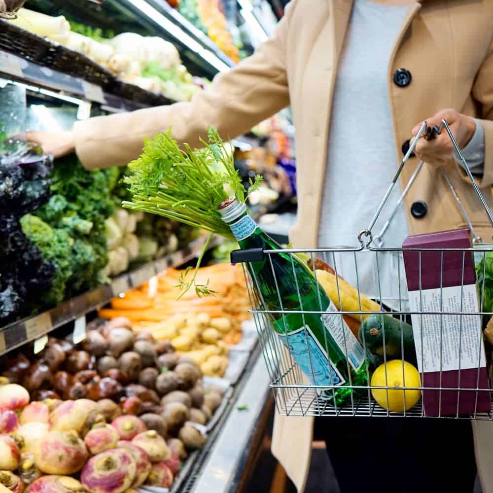 A woman reaching for a bunch of lettuce in a grocery store aisle. She is wearing a tan pea coat and holding a wire handbasket with some items inside.