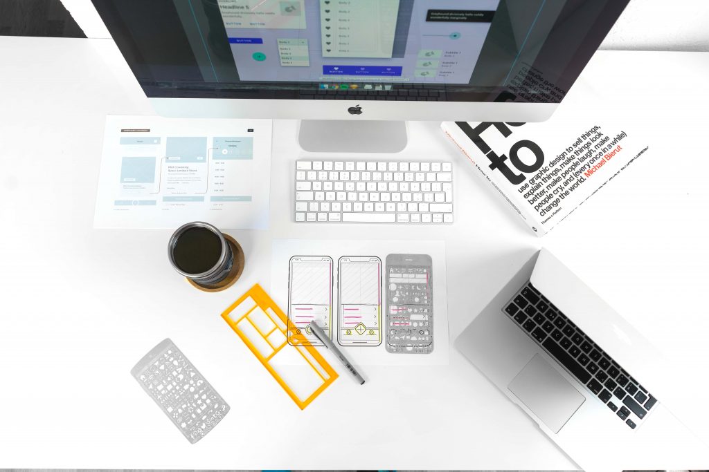 A desk with sketched designs for a mobile app. Also on the desk is a laptop, a desktop computer, a book about graphic design, and a cup of coffee.
