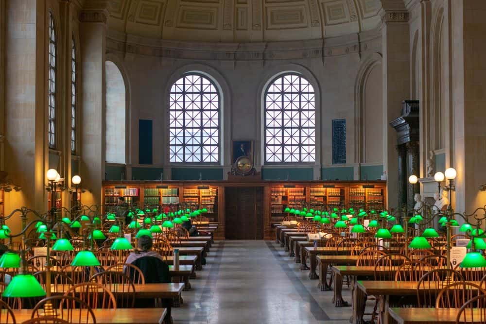 Bates Hall in the Boston Public Library. Empty long tables are lit by vintage green lamps. Two large windows are set in the far wall.