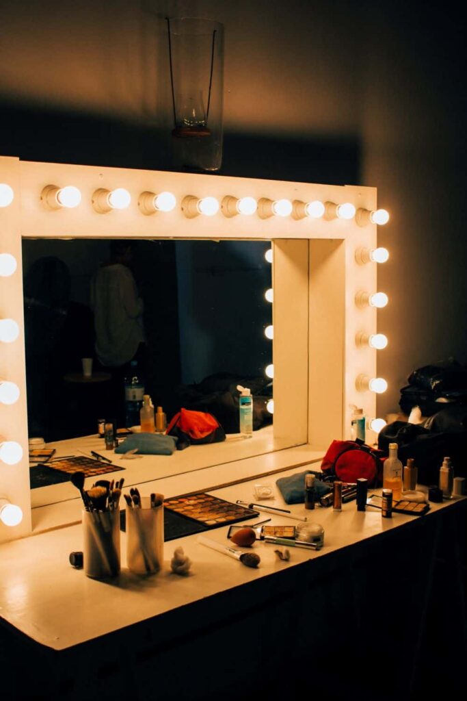 Large mirror lit around the border with light bulbs in a dressing room. Makeup covers the counter top