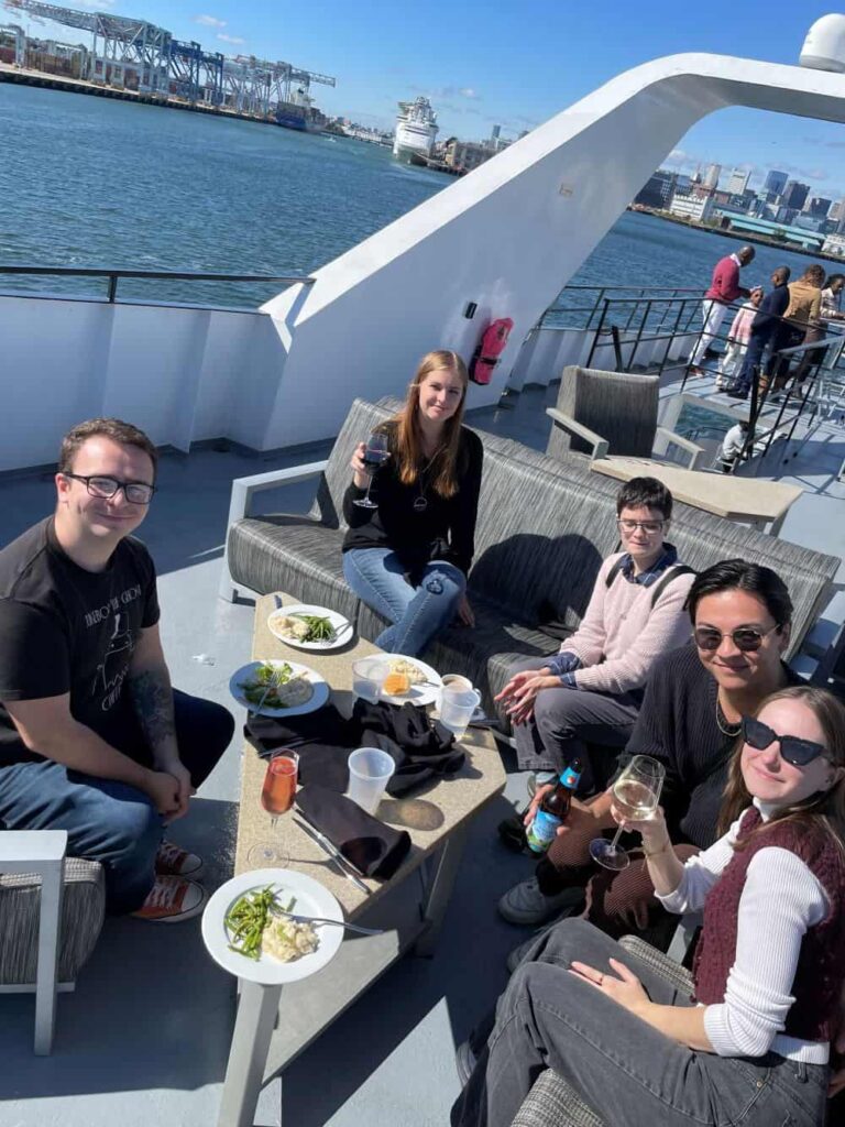 Graduate students gathered around a table on a cruise ship, enjoying food and beverages on a sunny afternoon