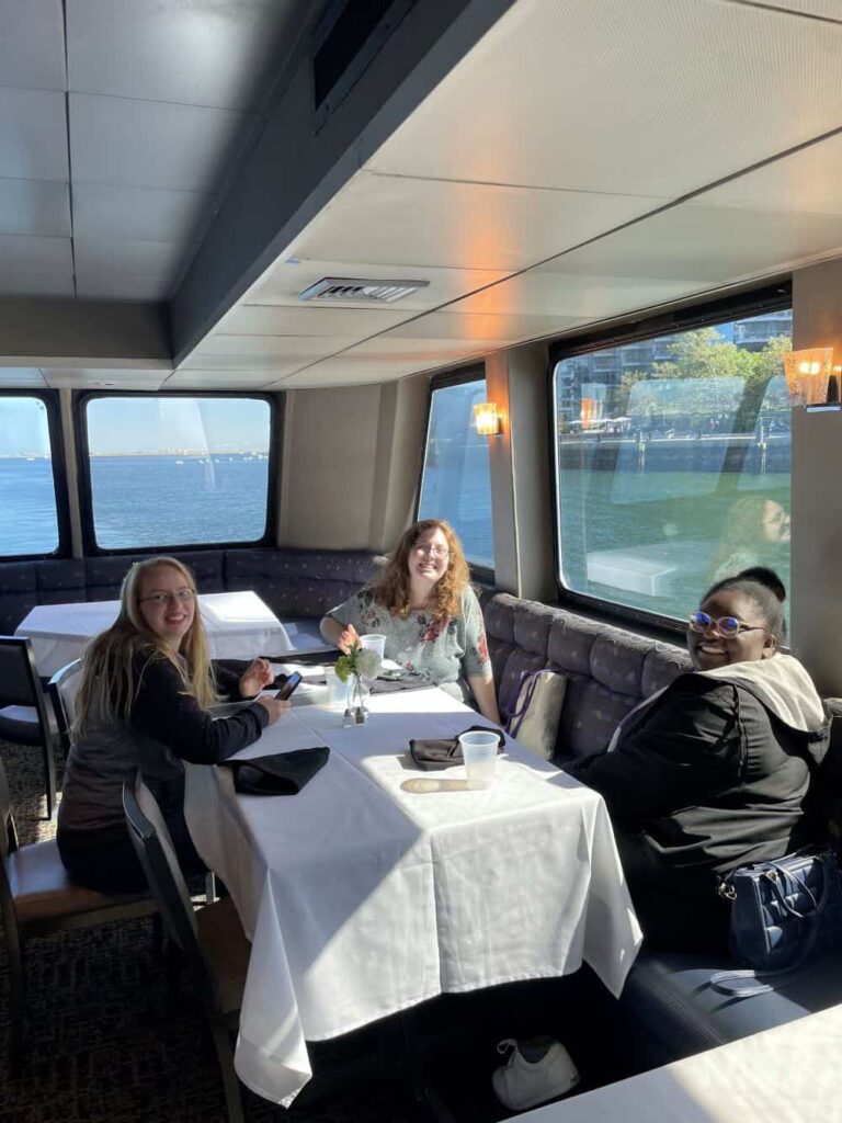 Three students gathered at a table on the lower level of a cruise ship for the GSA's Boston Harbor Lunch Cruise. They are smiling and having fun together