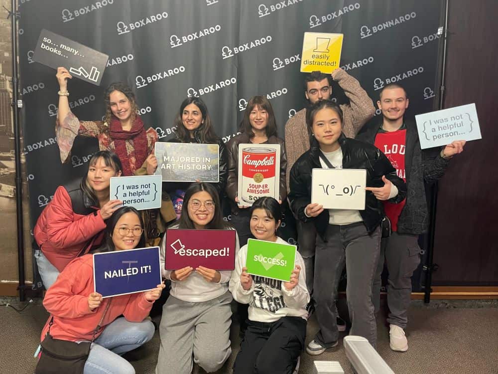 Ten students smiling posing in front of a "Boxaroo" backdrop after completing an escape room. They hold up signs with humorous captions like "easily distracted" and "I was not a helpful person....." 
