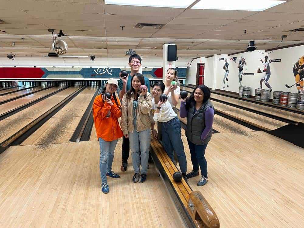 Six graduate students holding up bowling balls and smiling at the camera. They are in a Boston bowling alley for the IDGO bowling night