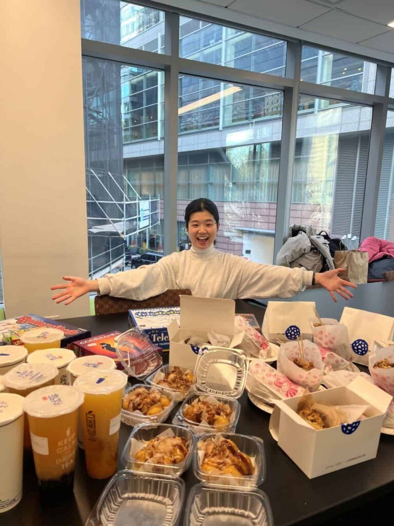 A graduate student in a white turtleneck  holds out her arms and smiles behind a table loaded with teas and deserts