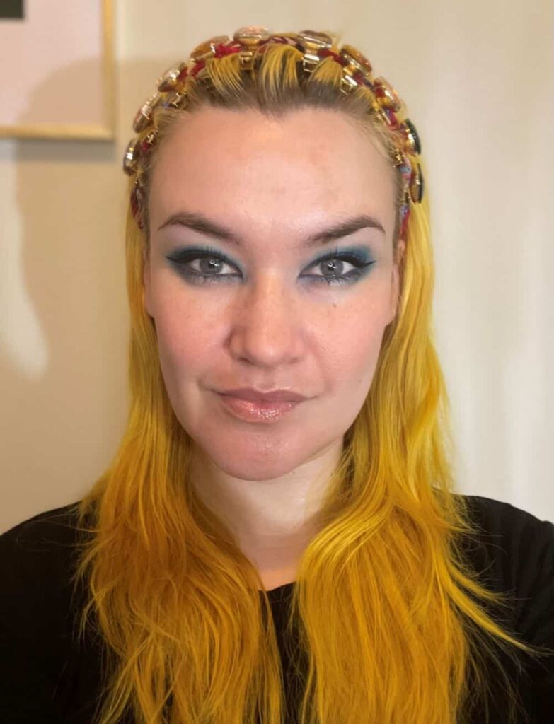 Sarah Burton, Editor-in-Chief of Page Turner Magazine, looks at the camera with a faint smile. Her hair is bright yellow and she wears a black top.