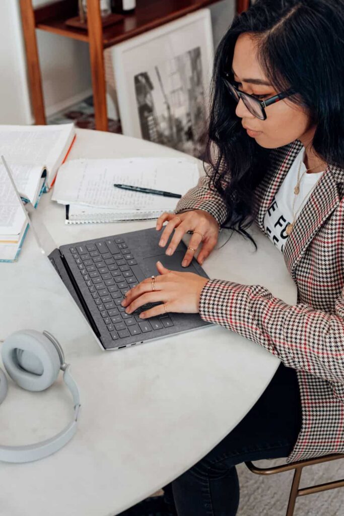 Fem-presenting person does research about salaries on a laptop. She wears a checkered blazer and glasses.