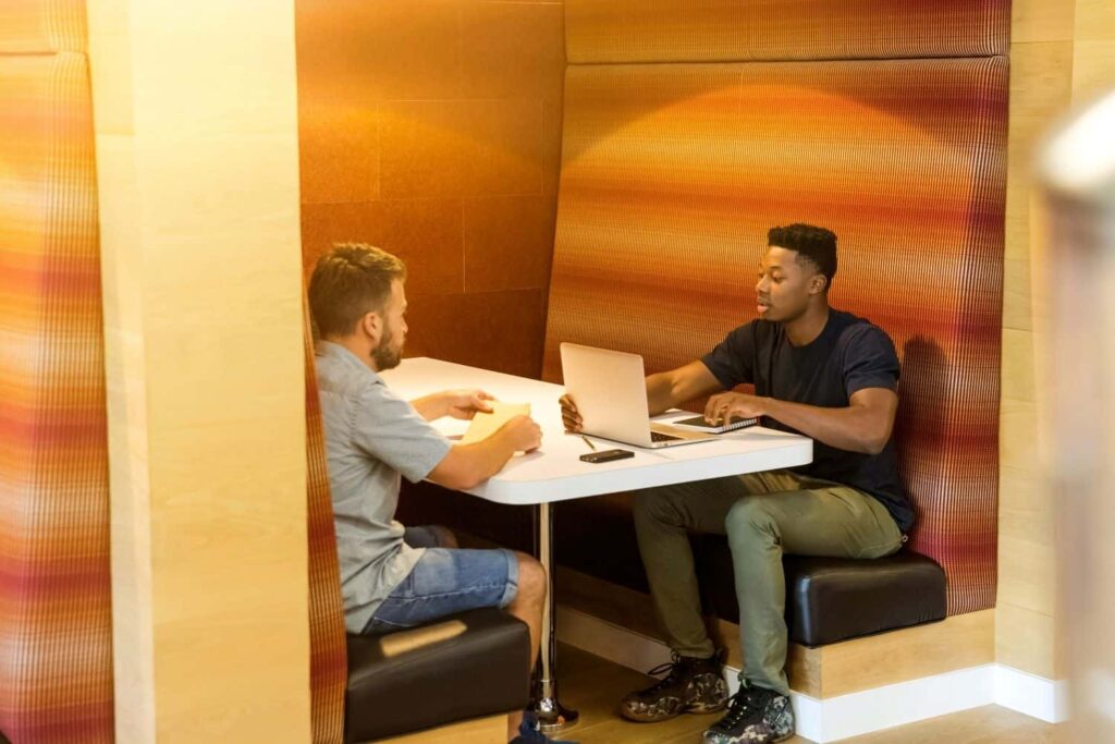 Two masc-presenting people sitting at a booth table. The person on the left looks at a paper, rehearsing his request for a raise. The person on the right is working on a laptop.