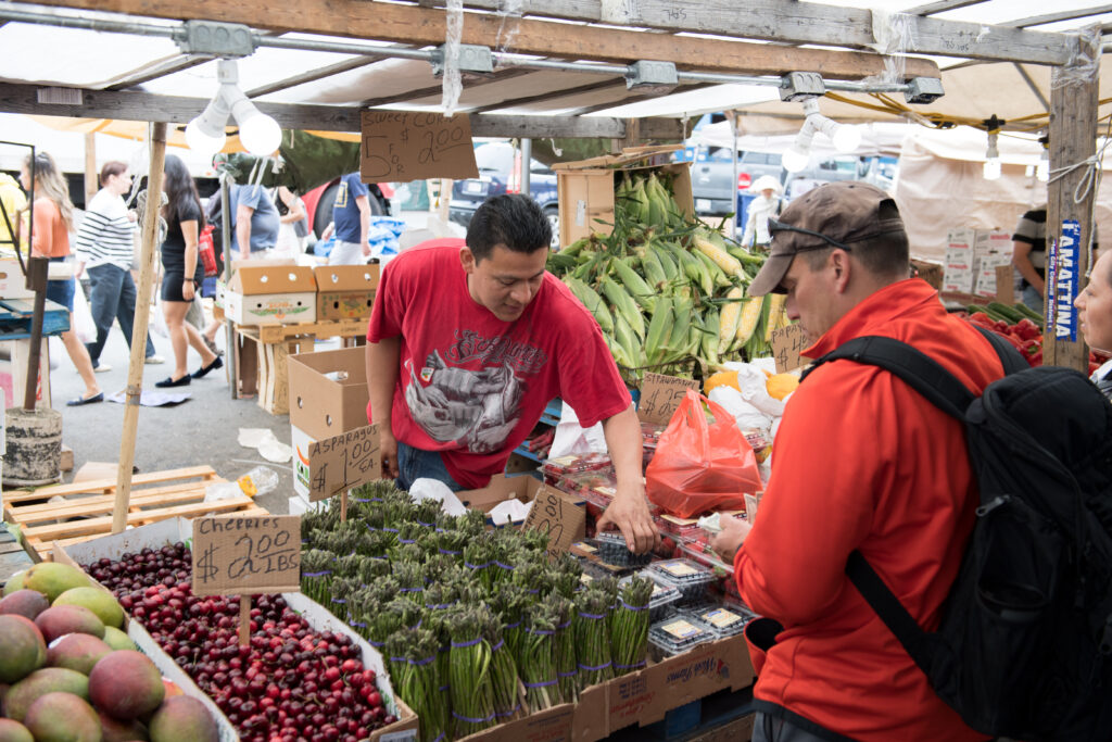 An open air tent selling mangoes, cherries, asparagus, and blueberries. A customer leans over to inspect some blueberries while talking with the vendor.