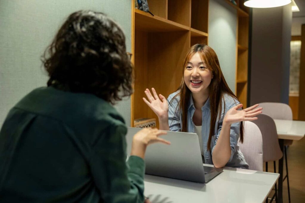 Two professionals sit at a table discussing something. The person facing the camera is smilimg and gesturing with open palms. She sits in front of a laptop and wears a blue button-up shirt. The person facing aaway from the camera has short curly hair and wears a green button-up.