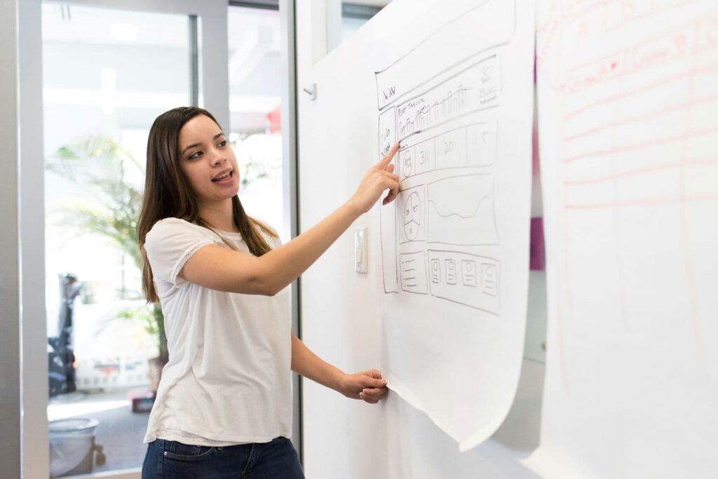 A senior marketing manager stands at a whiteboard, pointing at marketing data. She wears a white t-shirt and has long dark hair.